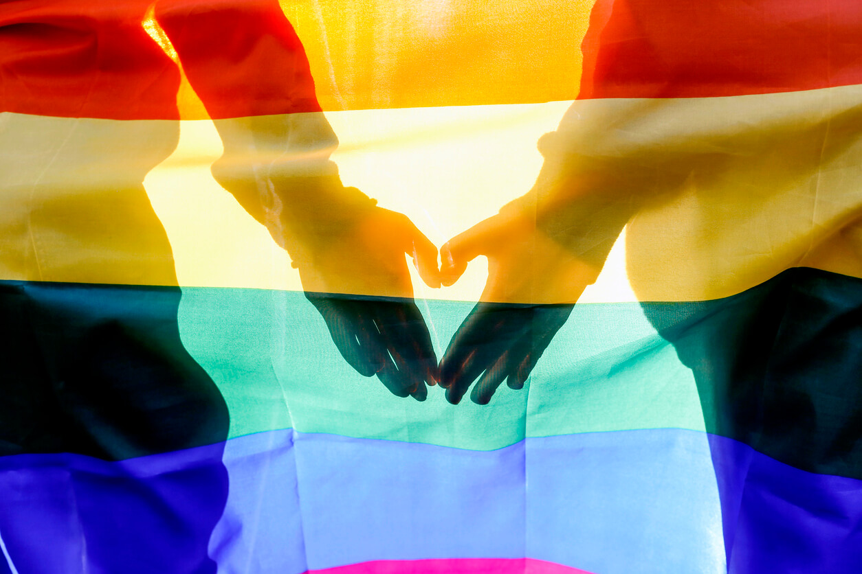 Two hands making the heart sign with the LGBTQ pride flag overlayed